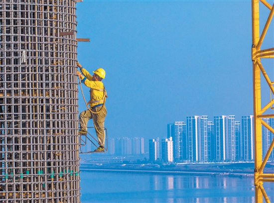 奮戰(zhàn)在百米索塔之上的建設(shè)者身影，與遠(yuǎn)處的高樓大廈遙呼相應(yīng)。