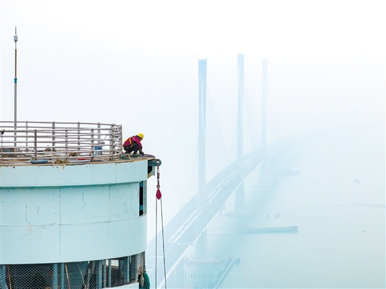 建設(shè)者在塔柱上施工，遠(yuǎn)處“海上長龍”若隱若現(xiàn)。